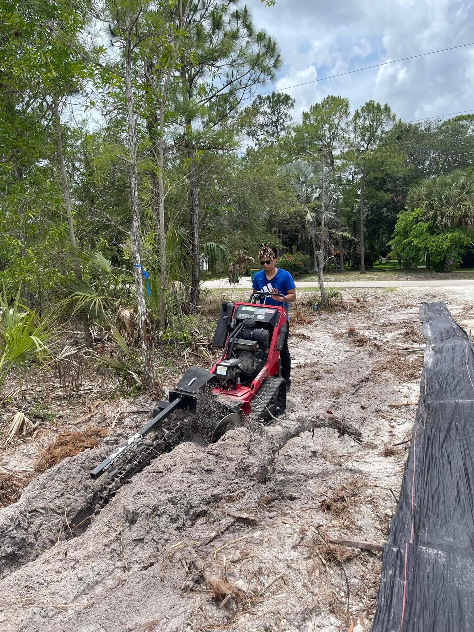 A man operates a red walk-behind trencher to dig a narrow trench in sandy soil.