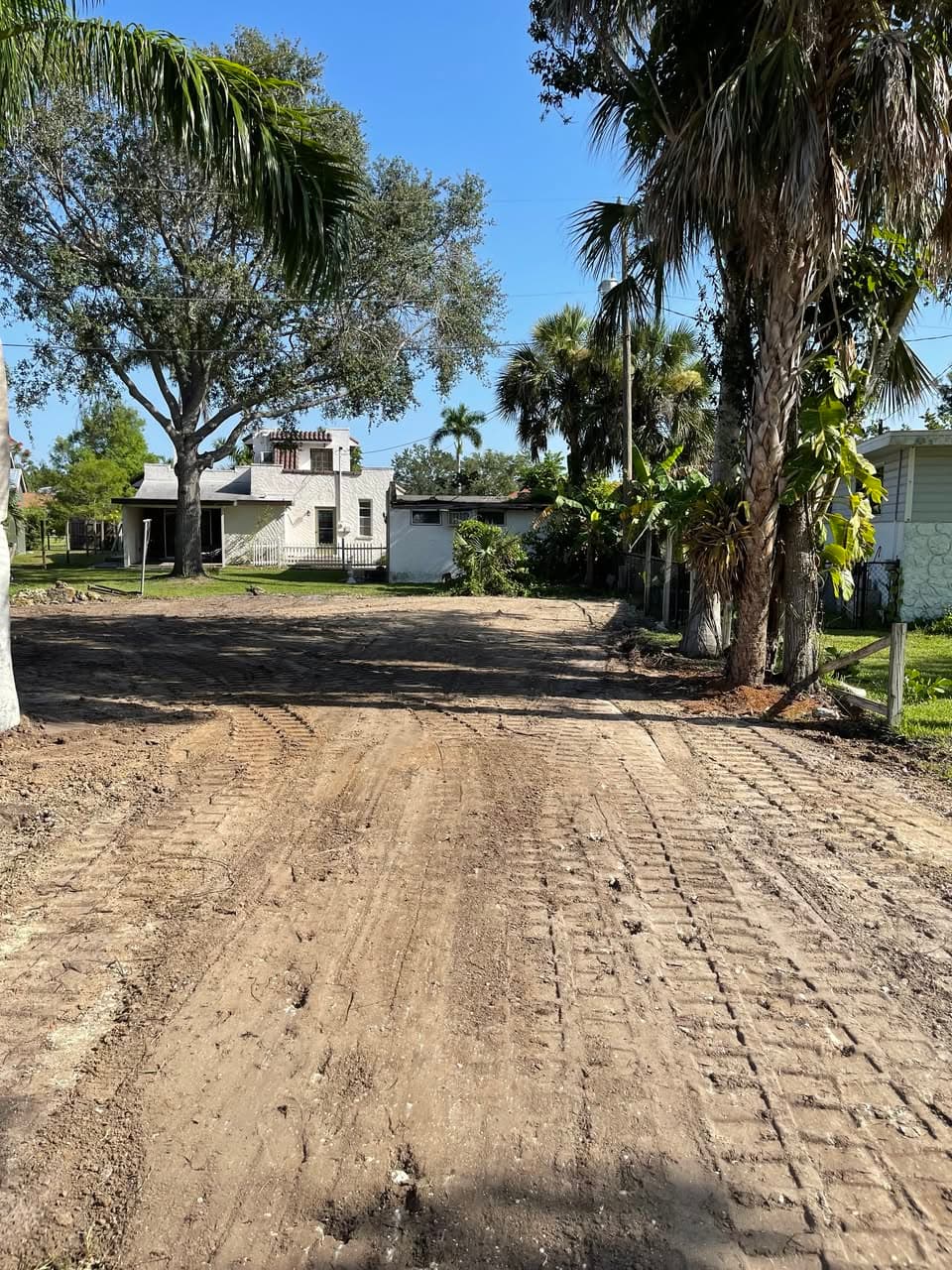 Dirt lot with heavy tread marks leading to a white house and palm trees.