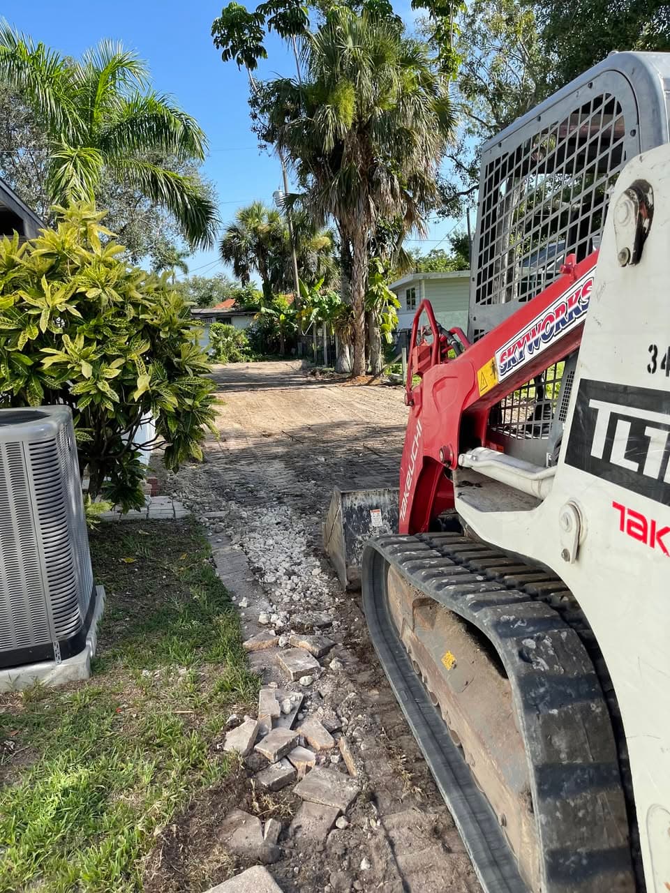 Red and white compact track loader on a dirt path in a tropical residential yard.
