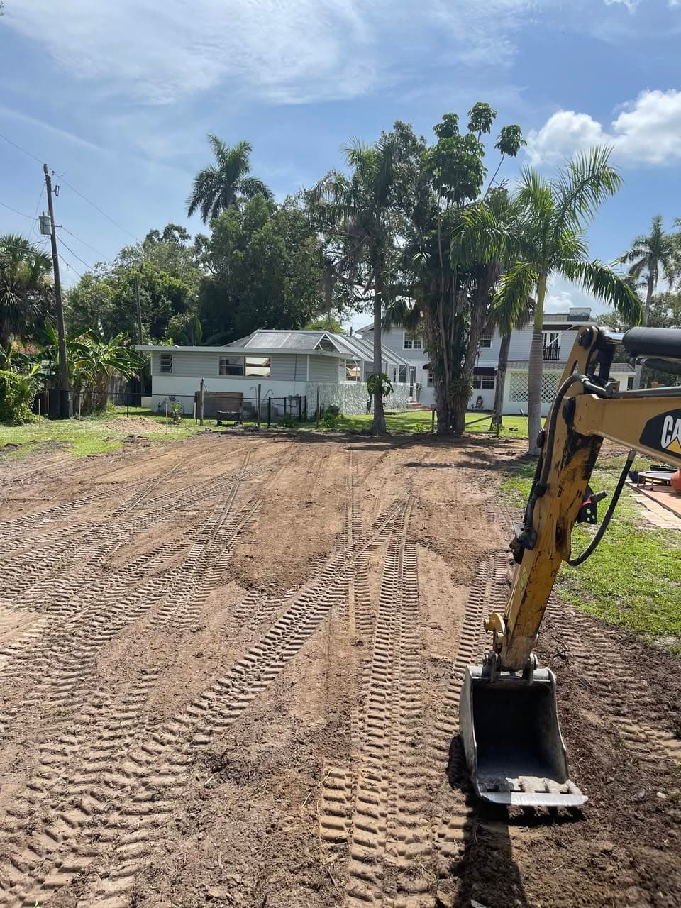 Yellow excavator bucket on a cleared dirt lot with tread marks in a tropical backyard.